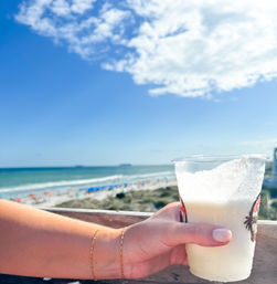 Hand with gold bracelets holding a frosty frozen tropical drink in a plastic cup on a sunny beach boardwalk overlooking turquoise ocean, white sand and blue sky