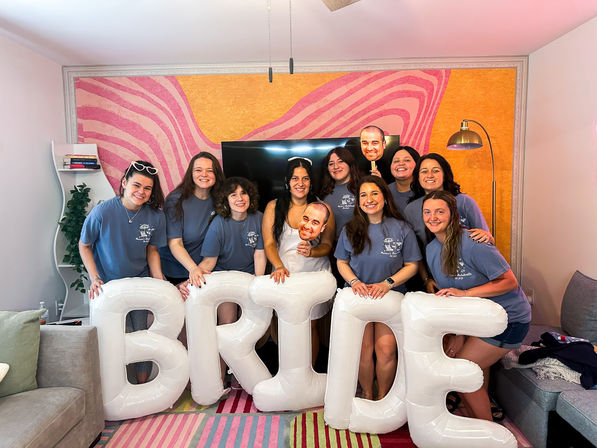 Smiling bridal party in matching blue shirts in a colorful living room holding oversized white inflatable letters spelling BRIDE, celebrating the bride-to-be in front of a pink swirled mural.