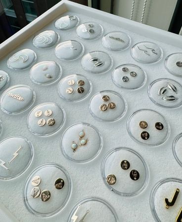 Jewelry studio display tray of round acrylic dishes on white felt, each holding small gold and silver charms, studs and opal connectors with shell, wave and lightning motifs.