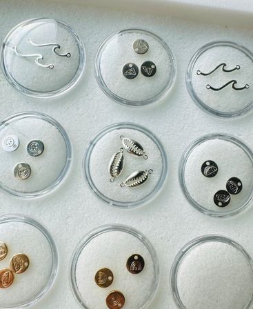Close-up flat lay of silver and gold jewelry charms—wave hooks, shell-style connectors and stamped round pendants—neatly arranged in clear circular dishes on white felt in a jewelry studio.