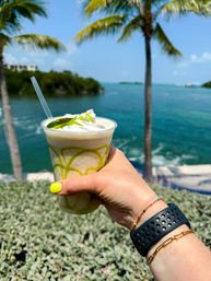 Hand with neon-yellow nail polish holding a lime-drizzled frozen drink topped with whipped cream and a lime wedge, set against turquoise ocean water, palm trees, and a sunny tropical shoreline.