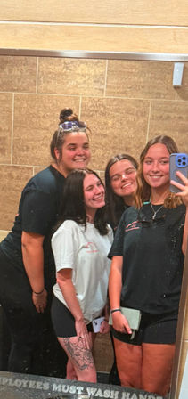 Four friends smiling for a mirror selfie in a tiled public restroom, wearing casual t-shirts and shorts; one has sunglasses on her head and a floral thigh tattoo, another holds a phone with a blue case, mirrored text 'Employees must wash hands' partially visible at the bottom.