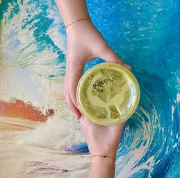 Overhead shot of two bracelet-adorned hands holding an iced matcha latte in a plastic cup with a handwritten name on the lid, set on a vibrant ocean-wave tabletop for a beachy cafe vibe.