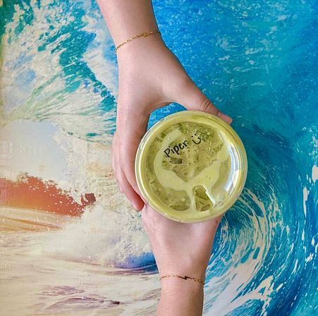 Overhead shot of two bracelet-adorned hands holding an iced matcha latte in a plastic cup with a handwritten name on the lid, set on a vibrant ocean-wave tabletop for a beachy cafe vibe.