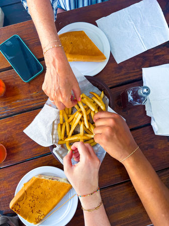 Top-down view of three hands reaching into a basket of golden French fries on a wooden picnic table, with sandwich halves on plates, napkins, a smartphone and a ketchup bottle — casual outdoor meal.