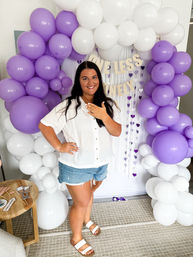 Smiling woman posing indoors under a purple-and-white balloon arch backdrop, showing an engagement ring while wearing a white shirt and denim shorts — celebratory engagement party photo.