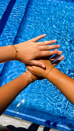 Close-up of three sun‑kissed hands stacked over sparkling blue swimming pool water, each wrist adorned with delicate gold bracelets — poolside summer friendship vibe.