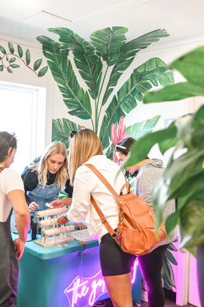 Shoppers examining bracelet display at a bright indoor pop-up shop with a large tropical leaf mural and pink neon sign