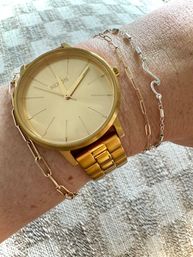 Close-up of a gold-tone minimalist analog wristwatch with a matching metal link band, paired with delicate gold and silver chain bracelets on a sunlit wrist over textured fabric background.