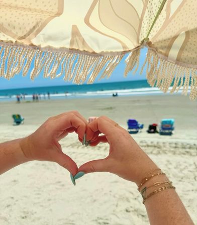 Heart-shaped hands with turquoise nails and layered bracelets under a fringed beach umbrella overlooking a sandy shore and turquoise ocean waves