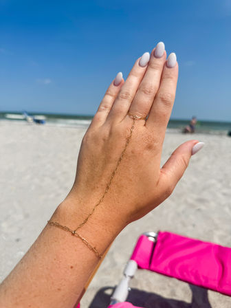 Close-up of a hand with white almond nails wearing a delicate gold hand-chain bracelet held up on a sunny beach with white sand, blue ocean waves and a pink beach chair in the background