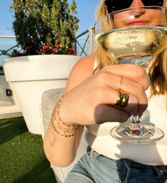 Close-up of a person toasting with a glass of white wine on a sunny rooftop patio, showing gold rings, stacked bracelets, sunglasses, white tank top and potted flowers in the background.