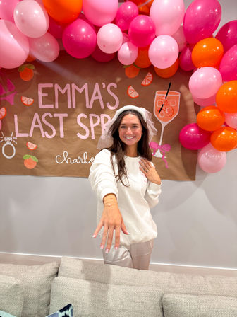 Beaming bride-to-be in a white sweatshirt and veil shows off her engagement ring at an indoor bachelorette celebration with pink and orange balloon garland and a hand-painted brown banner.