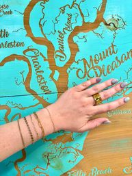 Hand wearing gold ring and delicate bracelets placed on a turquoise wooden map of Charleston area, featuring Mount Pleasant, Daniel Island and Folly Beach — coastal South Carolina decor.