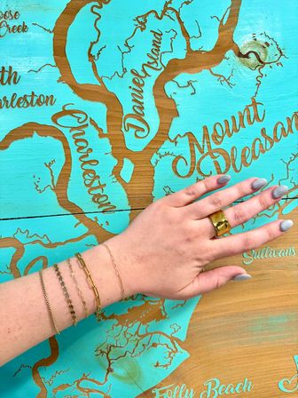 Hand wearing gold ring and delicate bracelets placed on a turquoise wooden map of Charleston area, featuring Mount Pleasant, Daniel Island and Folly Beach — coastal South Carolina decor.