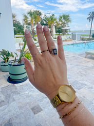 Hand with pale-pink manicure, dainty rings and gold watch held up by a pool at a tropical waterfront resort with palm trees, lounge chairs, and blue sky.