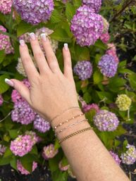 Close-up of a hand with white-tipped French manicure and layered gold and silver bracelets reaching toward pink and lavender hydrangea blooms in a garden