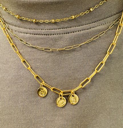Close-up of layered gold chain necklaces with three dainty round coin charms draped over a ribbed taupe crewneck top, fashion jewelry detail