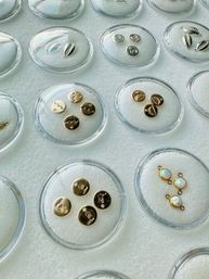 Close-up flatlay of gold coin-style charms, tiny shell and opal connectors arranged in clear round dishes on white felt — jewelry-making supplies display