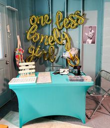 Indoor event table with teal tablecloth and gold foil balloons spelling "one less lonely girl" on a teal paneled wall, jewelry display trays on the table, clear folding chairs, a star-shaped balloon and a black-and-white male poster nearby.