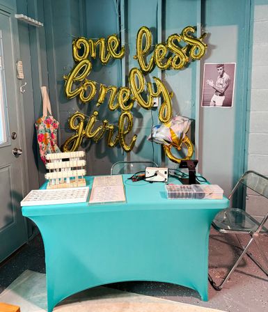 Indoor event table with teal tablecloth and gold foil balloons spelling "one less lonely girl" on a teal paneled wall, jewelry display trays on the table, clear folding chairs, a star-shaped balloon and a black-and-white male poster nearby.