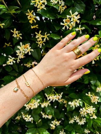 Close-up of a hand with neon yellow nails, chunky gold ring and delicate gold bracelets with small letter and padlock charms, posed against a green vine of white star-shaped flowers in a sunny garden