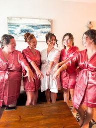 Bride in white robe and four bridesmaids in rose-pink satin robes smiling indoors on wedding morning, showing engagement rings in front of a coastal ocean painting.