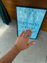 Hand with pale pink nails wearing a turquoise statement ring and delicate gold bracelets held over a concrete sidewalk in front of an aqua 'Permanent Jewelry' storefront sign with wood siding