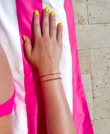 Sunlit poolside arm resting on a pink-and-white striped towel by a stone deck, neon yellow manicure and delicate gold and beaded bracelets visible