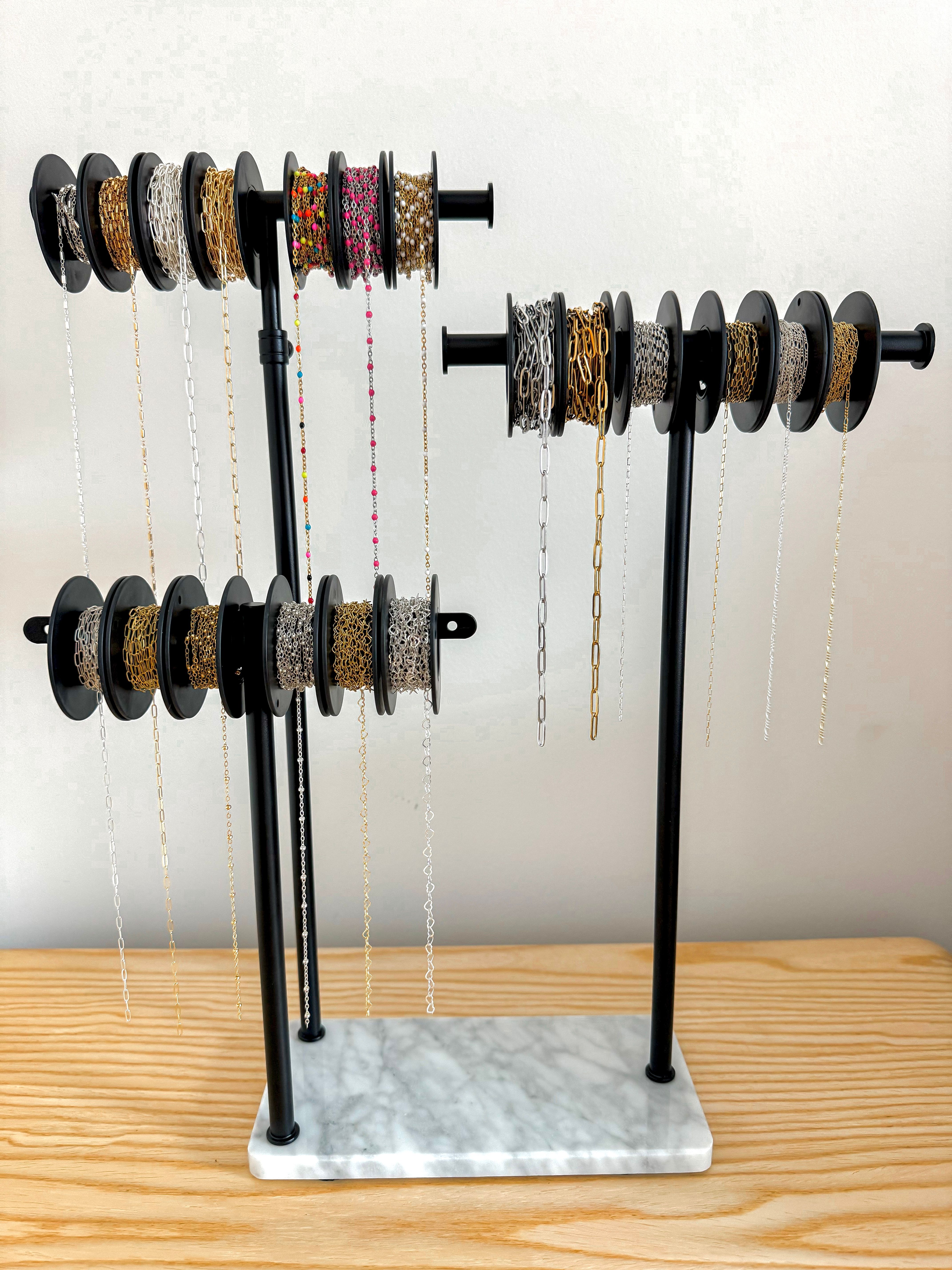 Black metal jewelry chain organizer with marble base on a wooden tabletop, multiple spools holding gold, silver and colorful beaded chains hanging against a pale wall.