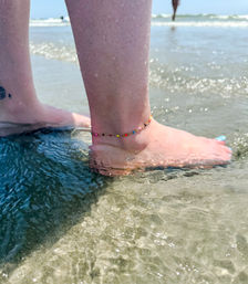 Close-up of bare feet wading in shallow ocean water on a sunny sandy beach, colorful beaded anklet and light-blue toenail polish adding a playful summer vibe.