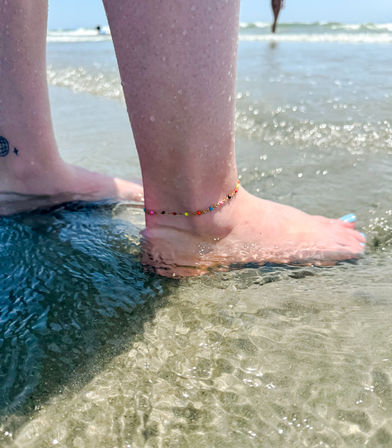 Close-up of bare feet wading in shallow ocean water on a sunny sandy beach, colorful beaded anklet and light-blue toenail polish adding a playful summer vibe.
