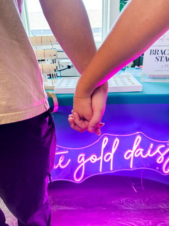 Close-up of two people holding hands at an indoor boutique counter, delicate bracelets on a wrist, pink-purple neon glow and jewelry display in the background