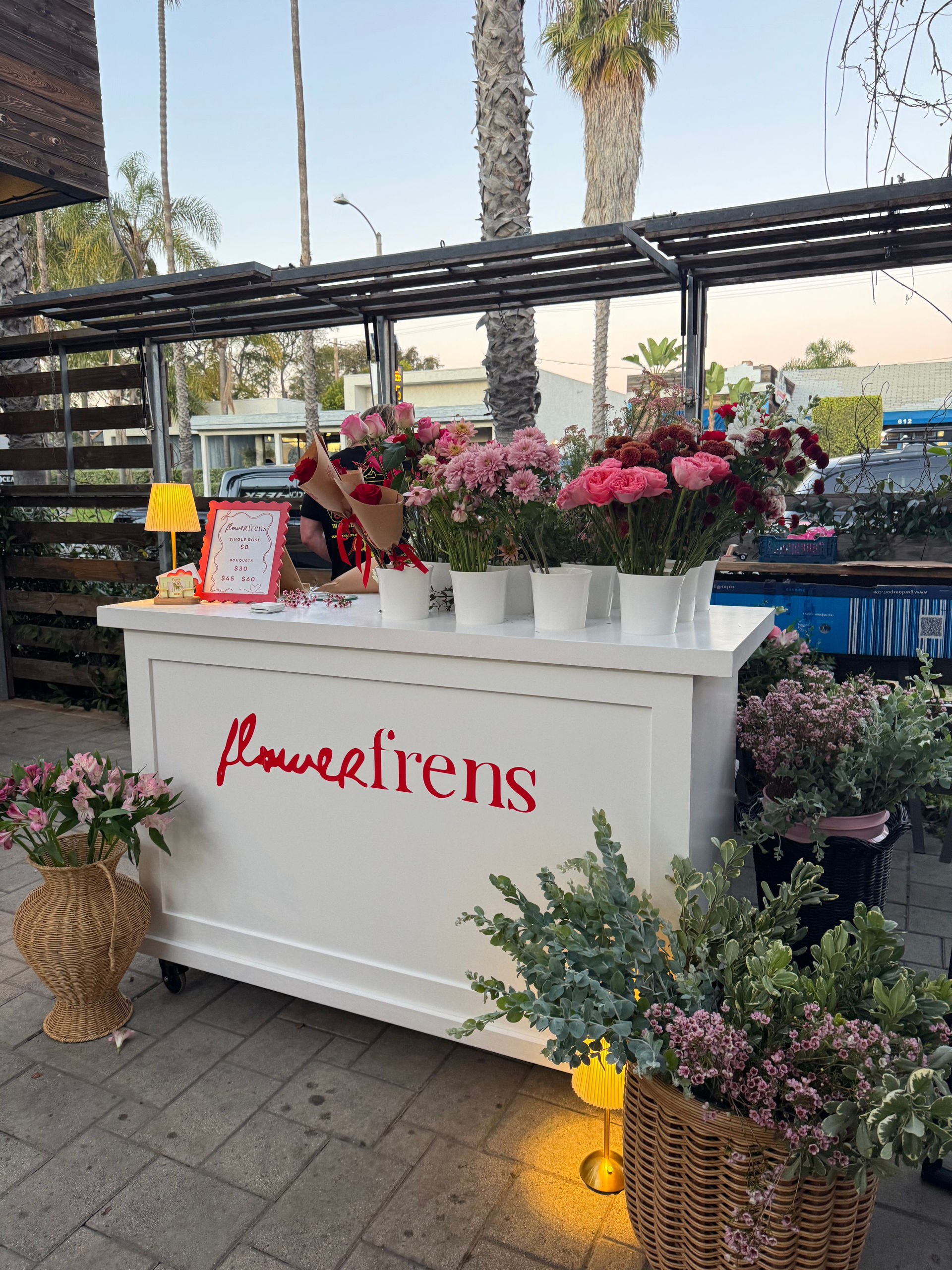 Outdoor white flower stand on a palm-lined sidewalk displaying pink roses, carnations and mixed bouquets in white pots and wicker baskets with warm lamp lighting