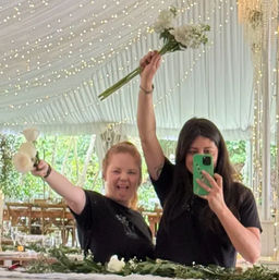 Two people in black shirts take a playful mirror selfie inside a white wedding tent draped with string lights, holding small white bouquets above a greenery-covered reception table.