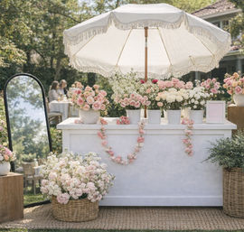 Outdoor garden flower stand with a white fringed umbrella — pastel pink and white roses and peonies in pots and wicker baskets, garlands draped across a white counter at a sunny wedding or garden party.
