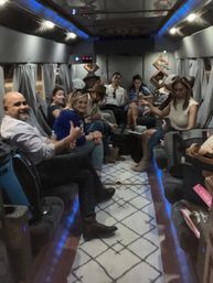 Group of adults socializing inside a party bus interior with blue LED accent lighting, cushioned bench seating along both sides, gray curtains, patterned runner rug and several people holding drinks.