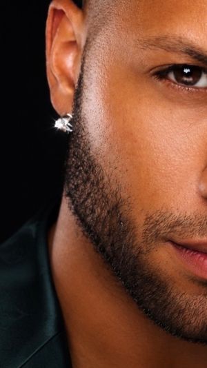 Close-up studio portrait of a man's chiseled jaw and trimmed beard, brown eye, glossy lips and a sparkling diamond stud earring against a black background.