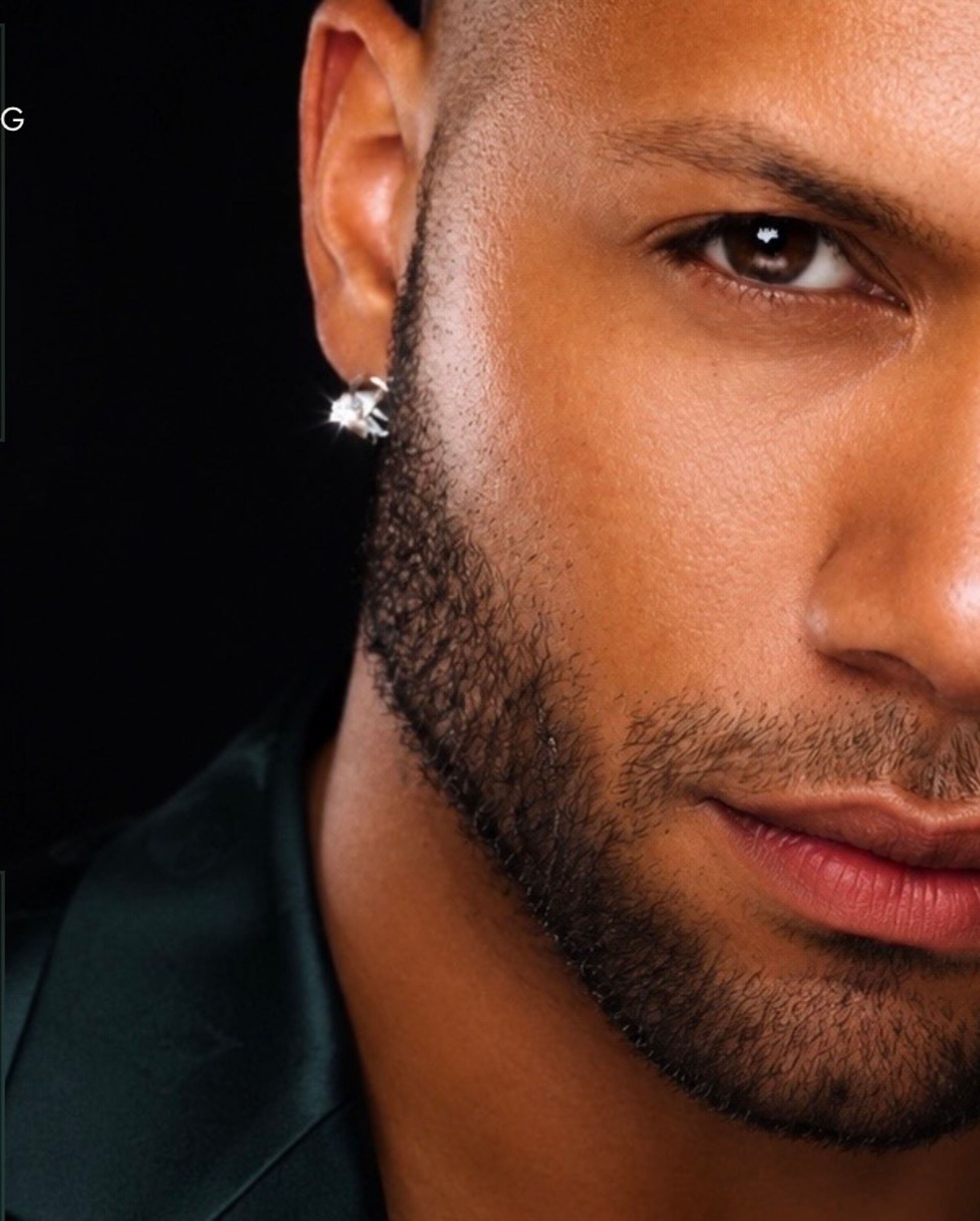 Close-up studio portrait of a man's chiseled jaw and trimmed beard, brown eye, glossy lips and a sparkling diamond stud earring against a black background.