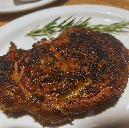 Close-up of a seasoned, charred grilled ribeye steak with a rosemary sprig on a white plate