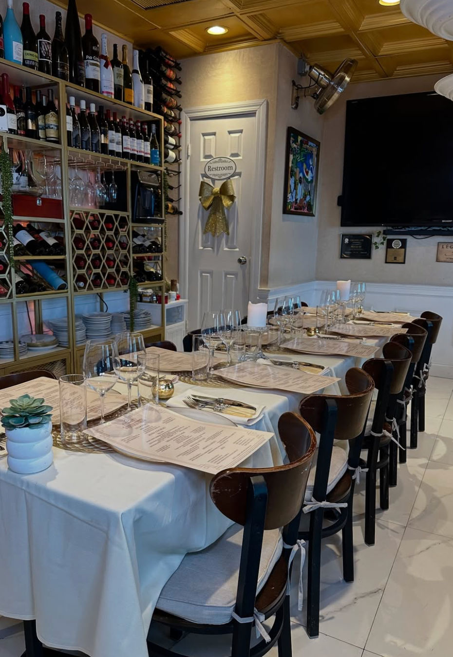 Cozy restaurant interior with a long white-cloth communal table set with menus, glassware and cutlery, cushioned wood chairs, and a wall of wine bottles and racks beside a restroom door with a gold bow.