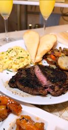 Steak-and-eggs brunch plate: pepper‑crusted medium‑rare steak, scrambled eggs with chives, roasted potatoes, toasted baguette halves, glazed shrimp appetizer and mimosas in champagne flutes.