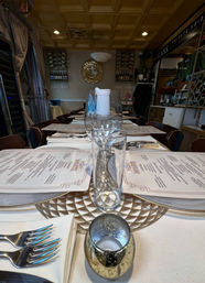 Intimate indoor restaurant table set for dinner with menus, stacked wine glasses holding a candle, silverware on napkins and wine bottles on shelves in the background.