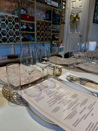 Elegant indoor restaurant table set for dinner with menus, multiple wine and water glasses, cutlery on a gold‑trimmed charger, honeycomb wine racks and bottles in the background, and a restroom door with a decorative bow.