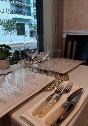 Elegant fine-dining restaurant table by a street-facing window with menus, two wine glasses and a water glass on a gold charger, pearl-handled cutlery on a folded napkin and a small potted plant.