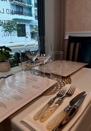 Elegant fine-dining restaurant table by a street-facing window with menus, two wine glasses and a water glass on a gold charger, pearl-handled cutlery on a folded napkin and a small potted plant.
