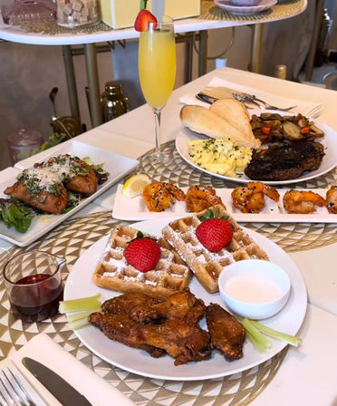 Bright brunch spread on a table: chicken-and-waffles dusted with powdered sugar and strawberries, celery and dipping sauce with syrup, glazed shrimp, steak and scrambled eggs with toast and mushrooms, a savory pastry on greens, and a strawberry-garnished mimosa.