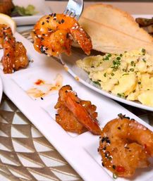 Sesame-glazed crispy shrimp appetizer on a long white plate, one on a fork, paired with chive mashed potatoes and toasted bread on a restaurant table