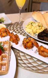 Brunch spread: crispy sesame-coated shrimp on a tasting tray, Belgian waffle with strawberries and powdered sugar, scrambled eggs, steak and biscuits, and a mimosa on a decorative table.