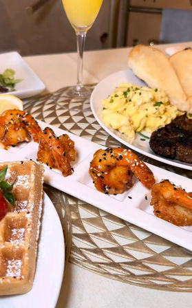 Brunch spread: crispy sesame-coated shrimp on a tasting tray, Belgian waffle with strawberries and powdered sugar, scrambled eggs, steak and biscuits, and a mimosa on a decorative table.